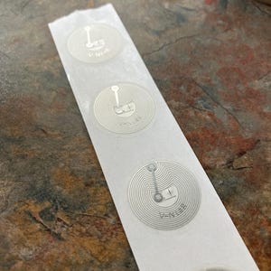 Pode incluir: Uma tira de etiquetas RFID prateadas com um design em espiral e o texto "V-N12B". As etiquetas estão em um suporte branco, com um fundo escuro.