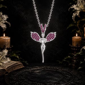 May include: A silver-toned pendant necklace featuring a stylized angel figure with outstretched wings. The wings and the bail are encrusted with numerous small, round, deep pink gemstones. The figure has a smooth, polished metallic finish.