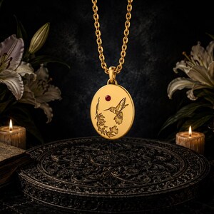 Op de afbeelding: Een goudkleurige ronde hanger aan een bijpassende ketting, voorzien van een gravure van een kolibrie bij bloemen en een kleine rode edelsteen. De hanger toont gedetailleerde lijntekeningen op een glad, goudkleurig oppervlak.