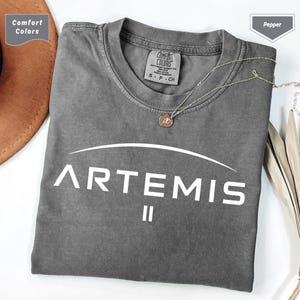 Può includere: Una maglietta grigia Comfort Colors con la scritta "ARTEMIS" in bianco, arcuata sopra il numero romano "II". Una collana d'argento con un ciondolo poggia sulla maglietta. La maglietta è piegata e un cappello marrone è parzialmente visibile.