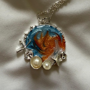 May include: A silver-toned pendant necklace featuring a circular resin charm with swirling blue, orange, and white colors. The charm is accented with two small silver starfish, two small silver seashells, and two round white pearls along the edge.