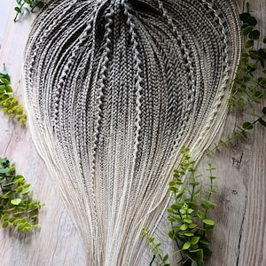May include: A set of synthetic hair extensions featuring numerous thin, tightly woven braids. The hair transitions in colour from a dark charcoal grey at the top roots to a light, pale blonde at the ends, creating a distinct ombre effect.