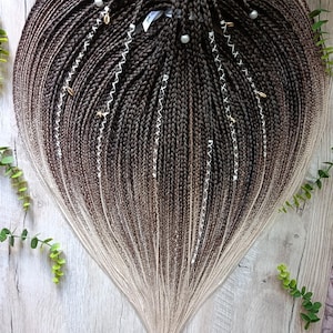 May include: A heart-shaped arrangement of dark brown to light blonde ombre synthetic hair braids. The braids feature white thread wraps, small gold-toned charms, and white spherical beads distributed throughout the hair strands.