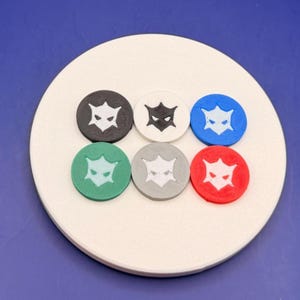 May include: Six circular tokens arranged in two rows on a round white base. Each token features a central cutout design of a stylized mask with pointed ears and narrow eyes. The tokens are black, white, blue, green, gray, and red in color.