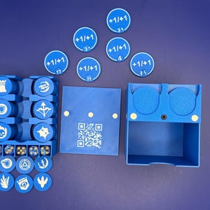 May include: A blue plastic game organizer set featuring a storage tray with slots for circular tokens, a separate lid with a printed QR code, and a matching box. The set includes multiple blue circular tokens marked with +1/+1 and various white icons.
