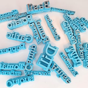 May include: A collection of light blue letter stamps with various names and words, including "Basil", "Rosemary", and "Tomatoes". The stamps are scattered across a white surface, showcasing their individual designs and text.