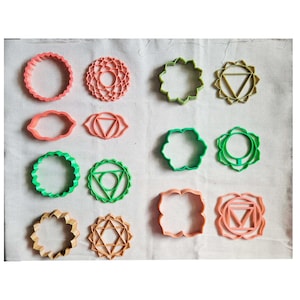 May include: A collection of colorful chakra-themed cookie cutters in various shapes, including circles, flower-like designs, and geometric patterns. The cutters are in shades of pink, green, and tan, arranged on a white surface.