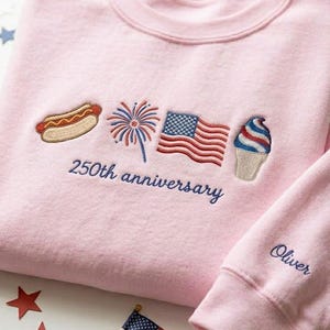 May include: A light pink sweatshirt with embroidered patriotic designs, including a hot dog, fireworks, an American flag, and an ice cream cone. The text "250th anniversary" is embroidered below the designs. The sleeve has the name "Oliver" embroidered.