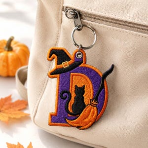 May include: An embroidered keychain featuring the letter D in purple and orange. The letter is decorated with a black witch hat, a black cat silhouette, and an orange broomstick. The keychain is attached to a silver metal ring and clasp.