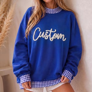 May include: A royal blue long-sleeve sweatshirt featuring a white, textured, cursive script applique that reads Custom across the chest. The garment includes a blue and white gingham patterned trim at the mock neckline, sleeve cuffs, and bottom hem.