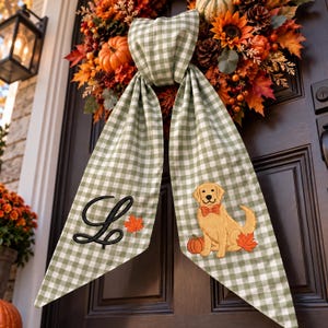May include: A sage green and white gingham fabric wreath sash featuring a black embroidered letter L, a small orange maple leaf, and an embroidered golden retriever wearing an orange bow tie next to a pumpkin and a maple leaf.