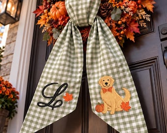 May include: A sage green and white gingham fabric wreath sash featuring a black embroidered letter L, a small orange maple leaf, and an embroidered golden retriever wearing an orange bow tie next to a pumpkin and a maple leaf.