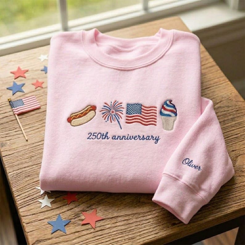 May include: A light pink sweatshirt with embroidered patriotic designs, including a hot dog, fireworks, an American flag, and an ice cream cone. The text "250th anniversary" is embroidered below the designs. The sleeve has the name "Oliver" embroidered.