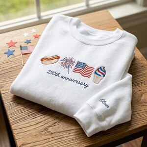 May include: White sweatshirt with embroidered patriotic icons: hot dog, fireworks, American flag, and ice cream cone. The text "250th anniversary" is embroidered below the icons. The sleeve cuff has the name "Oliver" embroidered in blue.
