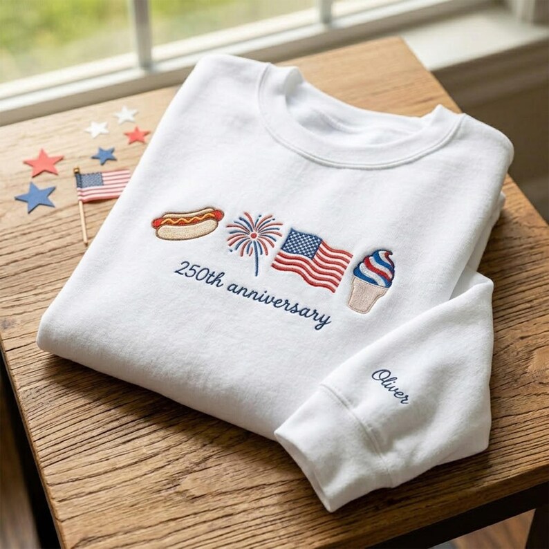 May include: White sweatshirt with embroidered patriotic icons: hot dog, fireworks, American flag, and ice cream cone. The text "250th anniversary" is embroidered below the icons. The sleeve cuff has the name "Oliver" embroidered in blue.
