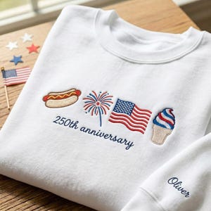 May include: White sweatshirt with embroidered patriotic designs, including a hot dog, fireworks, American flag, and ice cream cone. The text "250th anniversary" is embroidered below the designs. The sleeve has the name "Oliver" embroidered.