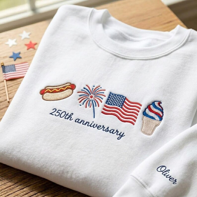 May include: White sweatshirt with embroidered patriotic designs, including a hot dog, fireworks, American flag, and ice cream cone. The text "250th anniversary" is embroidered below the designs. The sleeve has the name "Oliver" embroidered.