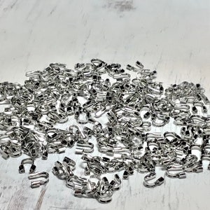 May include: A collection of small, shiny silver-colored metal findings, possibly jewelry components, scattered across a light-colored surface. The findings have a curved, open shape, suggesting they might be clasps or connectors for jewelry making.