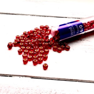 Puede incluir: Cuentas de semillas rojas con agujeros dorados, que se derraman de un tubo transparente. El tubo tiene una etiqueta azul y roja que dice "seed beads CZECH GLASS Round 6/0". Las cuentas están esparcidas sobre una superficie de madera blanca.