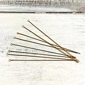 May include: A collection of gold-colored eye pins, each with a small loop at one end. The pins are arranged diagonally across a white, weathered wooden surface, creating a visually appealing pattern. The eye pins are used for jewelry making.