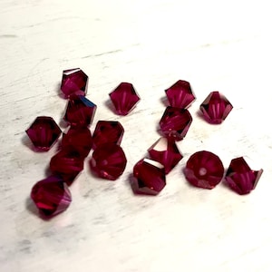 May include: A collection of faceted, dark red beads. The beads have multiple sides and a sparkling, translucent appearance. They are scattered on a white surface, creating a contrast that highlights their color and shape. These beads are suitable for jewelry making and crafting.