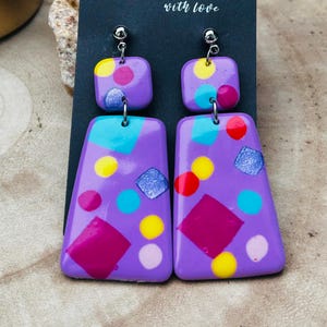 Peut inclure: Boucles d'oreilles violettes faites à la main avec des formes géométriques colorées et le texte "Handmade with love". Les boucles d'oreilles ont un dessus carré et un bas trapézoïdal, ornées d'accents roses, jaunes, bleus et scintillants.