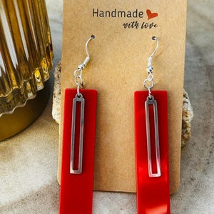 Può includere: Un paio di orecchini rettangolari rossi con un accento rettangolare argentato. Gli orecchini sono esposti su una carta marrone con la scritta "Handmade with love". Gli orecchini hanno ganci argentati.