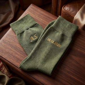 May include: A pair of olive green socks with gold embroidered details. One sock features the initials "AS", while the other displays the date "06.13.2026". The socks are displayed on a dark wooden surface.