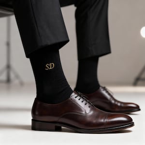 Può includere: Scarpe eleganti in pelle marrone con lacci neri. I calzini neri sono ricamati con le lettere dorate "SD". La persona indossa pantaloni neri. Stile classico.