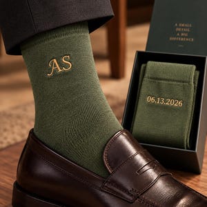 Puede incluir: Calcetines de color verde oliva con las iniciales AS bordadas en dorado en el tobillo. Un par a juego se encuentra dentro de una caja de regalo verde oscuro con el texto A SMALL DETAIL A BIG DIFFERENCE y la fecha 06.13.2026 bordados en oro.