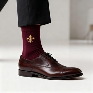 May include: Burgundy dress socks with a gold fleur-de-lis emblem, paired with brown leather dress shoes. The socks are visible beneath black dress pants, creating a classic and stylish look.