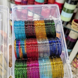 May include: A clear plastic container filled with stacks of colorful glass bangles. The bangles are in various colors, including blue, red, green, yellow, and purple. The text "VLUXE FASHION" is in the upper right corner.