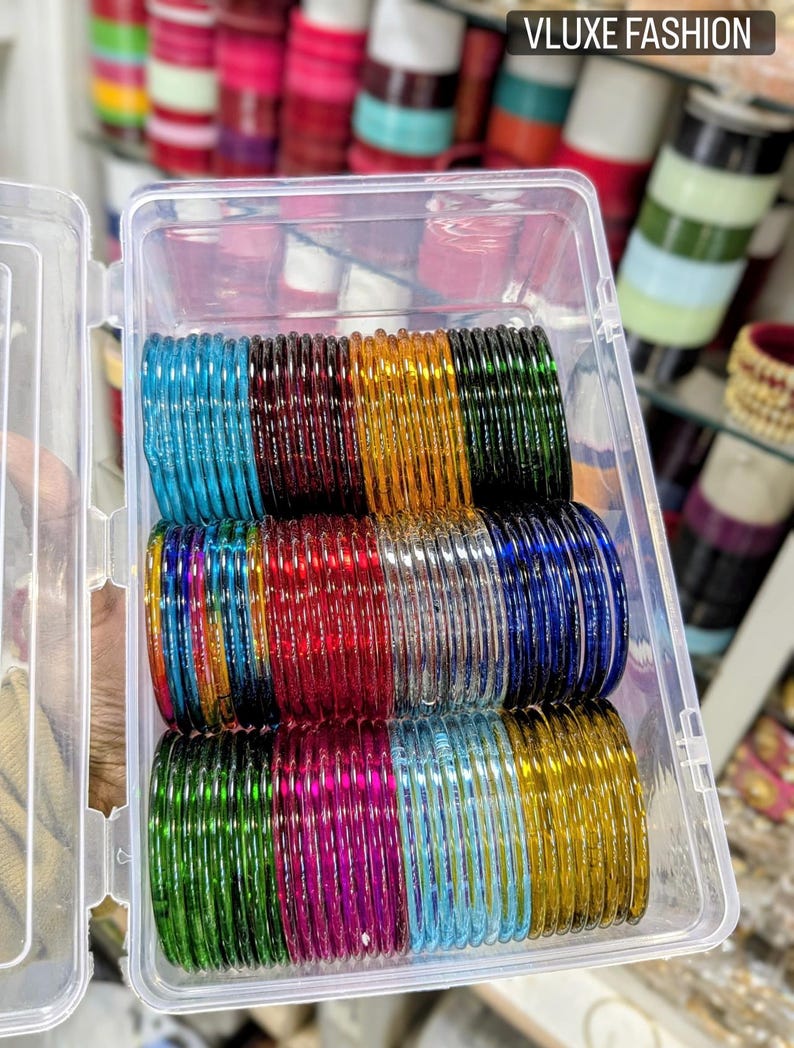 May include: A clear plastic container filled with stacks of colorful glass bangles. The bangles are in various colors, including blue, red, green, yellow, and purple. The text "VLUXE FASHION" is in the upper right corner.