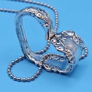 May include: A silver necklace with a pendant made from a repurposed spoon. The pendant has decorative floral detailing and hangs from a beaded chain. The background is a light blue.