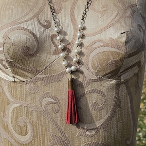May include: A long necklace with faux pearls and a red tassel pendant. The necklace has a dark chain and is displayed on a mannequin with a patterned fabric.