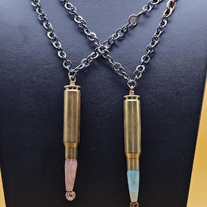 May include: Two bullet-shaped pendants with gemstone tips hang from gunmetal chains. One pendant has a pink gemstone, the other a blue gemstone. The necklaces are displayed on a black stand against a yellow background.