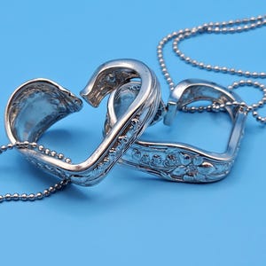 May include: A silver heart-shaped pendant necklace crafted from repurposed silverware. The pendant features two interlocking heart halves, each with ornate floral detailing. The necklace hangs on a silver ball chain against a blue backdrop.