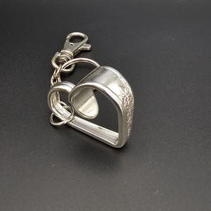 May include: A silver-coloured keyring featuring a heart-shaped design made from repurposed silverware. The keyring includes a clasp and a ring for attaching keys. The heart shape is formed by the curved handles of the silverware.