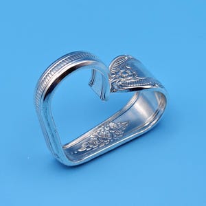 May include: A silver napkin ring in the shape of a heart, crafted from a repurposed spoon. The ring features decorative floral patterns and a ridged edge, set against a solid blue background. A unique and charming table accessory.