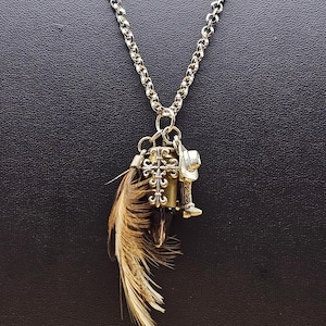 May include: Silver-toned necklace with a chain link design. The pendant features a cross, a small cowboy hat, a boot, and a feather. The necklace is displayed against a dark background.