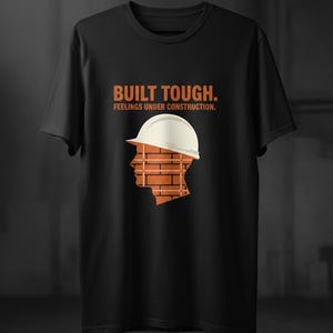 May include: Black t-shirt with the text "BUILT TOUGH. FEELINGS UNDER CONSTRUCTION." in orange. The shirt features a graphic of a head wearing a white hard hat, with the head's silhouette filled with a brick pattern.
