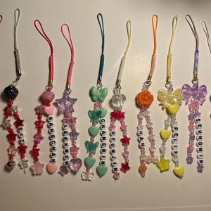 以下が含まれることがあります： さまざまな色のビーズの携帯電話チャームのコレクション。各チャームは、星、ハート、リボンなど、さまざまなデザインが特徴で、白い文字ビーズで名前が綴られています。チャームはカラフルなコードに取り付けられています。
