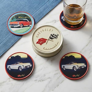 May include: Set of vintage car coasters with a red trim. The coasters feature classic car illustrations and a tin with the Chevrolet logo and checkered flags. A glass of amber liquid sits on one coaster.