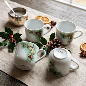 May include: Four white ceramic mugs featuring a hand-painted design of green pine needles and red holly berries. Two mugs are upright, while two are inverted, revealing the signature N. Ballar 1992 on the base of the mugs in black ink.