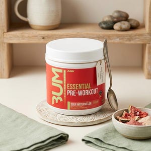 May include: A white plastic tub of BUM Essential Pre-Workout powder in Sour Watermelon flavor. The label is red with white and yellow text. A silver spoon rests against the side of the tub, which sits on a round, textured, off-white stone coaster.