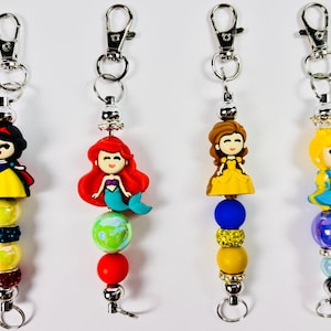 May include: Four keychains featuring cartoon princess figures. Each keychain has a silver clasp, decorative beads in various colors, and a princess figurine. The princesses include Snow White, Ariel, Belle, and Cinderella.