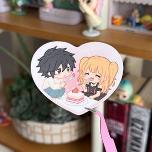 May include: A heart-shaped sticker featuring a chibi-style illustration of a dark-haired character and a blonde character eating a strawberry shortcake together. The characters are drawn with simple lines and soft colours, set against a pale pink background.