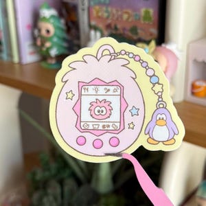 May include: A die-cut sticker featuring a light pink, egg-shaped digital pet device with a screen displaying a small pink creature. The device has three circular buttons and is attached to a beaded chain with a small purple penguin charm.