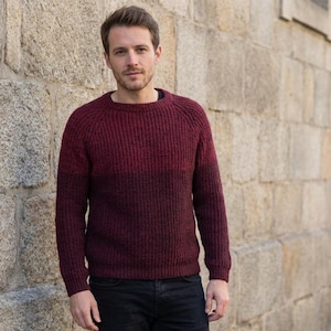 May include: A burgundy crew neck sweater with a ribbed texture. The sweater has long sleeves and is worn with black pants. The person is standing in front of a stone wall.