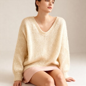 May include: A cream-colored, oversized sweater with a deep V-neck. The sweater has long sleeves and a loose knit texture. The model is wearing a pink skirt. The sweater appears soft and comfortable, suitable for casual wear.